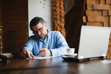 Erkek serbest yazar yeni bir iş projesi için bir kafede çalışıyor. Masadaki büyük bir pencerede oturuyor. Bir fincan kahveyle dizüstü bilgisayara bakıyor..