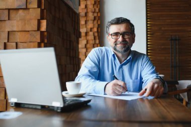 Kafede kahve içen olgun bir iş adamı. Güzel gözlükler takan, dizüstü bilgisayar kullanan, kameraya bakıp gülümseyen yakışıklı bir adamın portresi. Kahve molası.