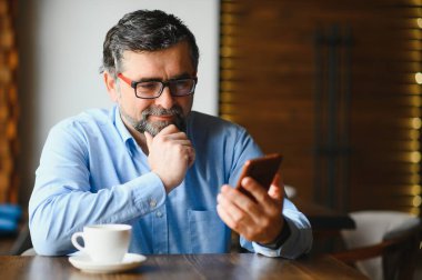 Restoranda oturup öğle yemeğini bekleyen mutlu iş adamı. Akıllı telefon kullanıyor ve biriyle konuşuyor. İş dünyası yaşlıları yaşam tarzı konsepti