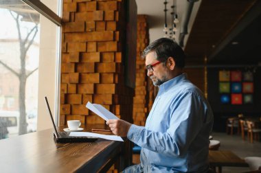 Erkek serbest yazar yeni bir iş projesi için bir kafede çalışıyor. Masadaki büyük bir pencerede oturuyor. Bir fincan kahveyle dizüstü bilgisayara bakıyor..
