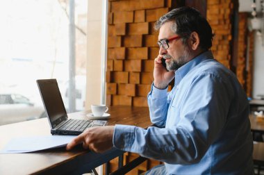 Restoranda oturup öğle yemeğini bekleyen mutlu iş adamı. Akıllı telefon kullanıyor ve biriyle konuşuyor. İş dünyası yaşlıları yaşam tarzı konsepti