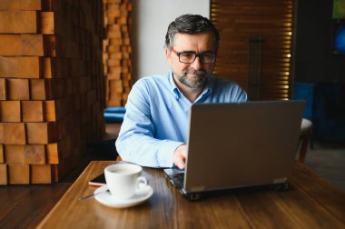 Kafede kahve içen olgun bir iş adamı. Güzel gözlükler takan, dizüstü bilgisayar kullanan, kameraya bakıp gülümseyen yakışıklı bir adamın portresi. Kahve molası.