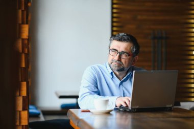 İş, teknoloji ve insan kavramı, dizüstü bilgisayarlı kıdemli işadamı modern kafede kahve içiyor.