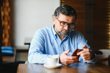 Restoranda oturup öğle yemeğini bekleyen mutlu iş adamı. Akıllı telefon kullanıyor ve biriyle konuşuyor. İş dünyası yaşlıları yaşam tarzı konsepti