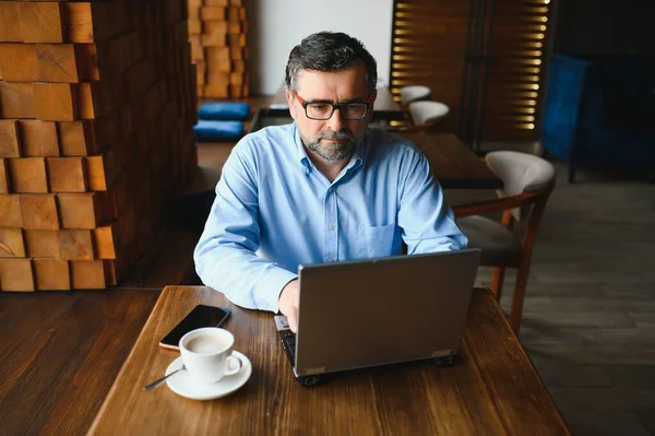 İş, teknoloji ve insan kavramı, dizüstü bilgisayarlı kıdemli işadamı modern kafede kahve içiyor.