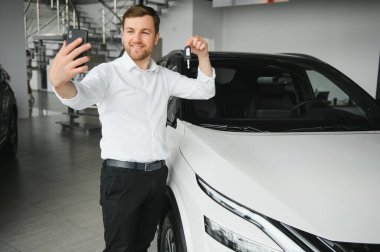 Genç adam yeni bir araç içinde araba Bayilik seçimi ve Smartphone'da fotoğraf yapma