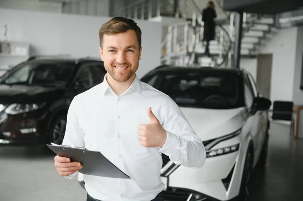 portrait of open-minded professional salesman in cars showroom, caucasian man in white formal shirt stands next to luxurious car and looks at camera.