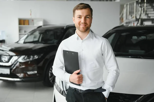 Assistant in vehicle search. Portrait of a handsome young car sales man in formalwear holding a clipboard and looking at camera in a car dealership.