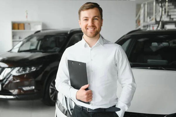 portrait of open-minded professional salesman in cars showroom, caucasian man in white formal shirt stands next to luxurious car and looks at camera.