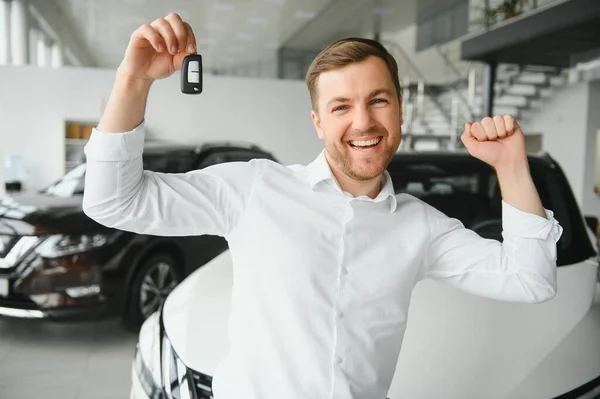 Happy buyer holding keys near the car in front of the modern avtosalon building.