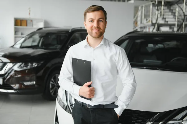 portrait of open-minded professional salesman in cars showroom, caucasian man in white formal shirt stands next to luxurious car and looks at camera.