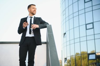 Molada resmi takım elbiseli sakallı iş adamı cep telefonu kullanıyor. Şehir merkezindeki modern şehir sokaklarının arka planında elinde kahve fincanıyla duran bir iş adamı. Boşluğu kopyala.