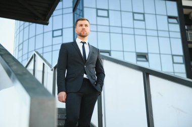 Modern iş adamı. Kendine güvenen, takım elbiseli genç bir adam arka planda şehir manzarasıyla dışarıda dikilirken gözlerini kaçırıyor..