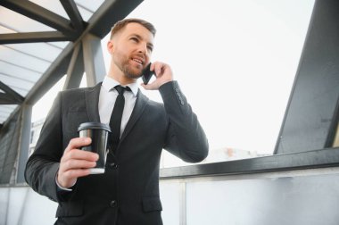 Molada resmi takım elbiseli sakallı iş adamı cep telefonu kullanıyor. Şehir merkezindeki modern şehir sokaklarının arka planında elinde kahve fincanıyla duran bir iş adamı. Boşluğu kopyala.