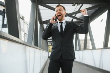 Mutlu işadamı gülümseyen siyah takım elbise ve modern smartphone kullanma office sabah erken, gece gökdelen ofisinin dururken anlaşmak başarılı işveren, parıltılı ışık.