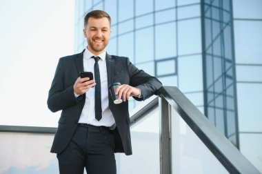 Molada resmi takım elbiseli sakallı iş adamı cep telefonu kullanıyor. Şehir merkezindeki modern şehir sokaklarının arka planında elinde kahve fincanıyla duran bir iş adamı. Boşluğu kopyala.