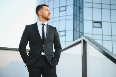 Modern iş adamı. Kendine güvenen, takım elbiseli genç bir adam arka planda şehir manzarasıyla dışarıda dikilirken gözlerini kaçırıyor..