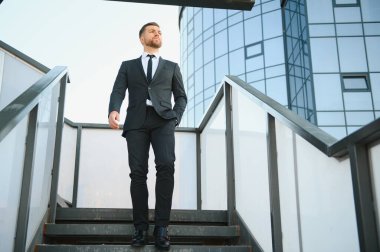 Modern iş adamı. Kendine güvenen, takım elbiseli genç bir adam arka planda şehir manzarasıyla dışarıda dikilirken gözlerini kaçırıyor..