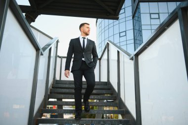 Modern iş adamı. Kendine güvenen, takım elbiseli genç bir adam arka planda şehir manzarasıyla dışarıda dikilirken gözlerini kaçırıyor..