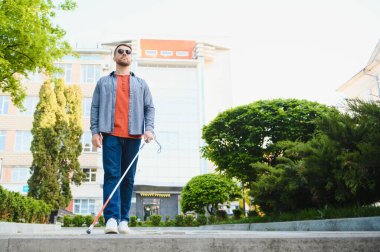 Şehirde sokakta yürüyen beyaz bastonlu genç kör adam.