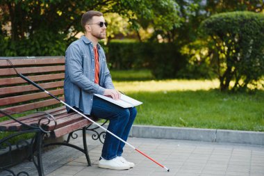 Blind man reading book on bench in park. clipart