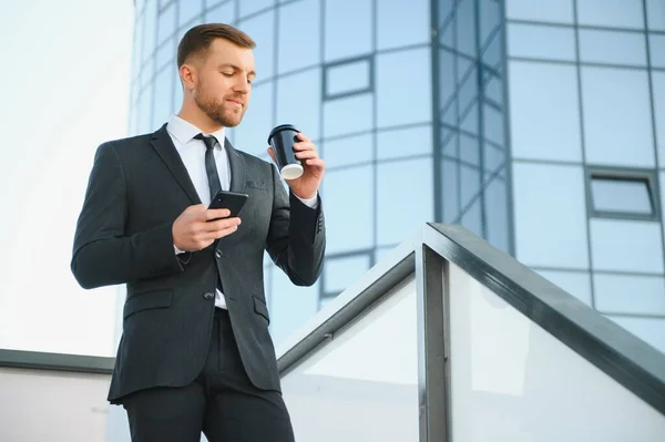 Bearded businessman in formal suit on break using mobile phone use smartphone. business man standing outside on modern urban city street background with coffee cup in downtown outdoors. copy space.