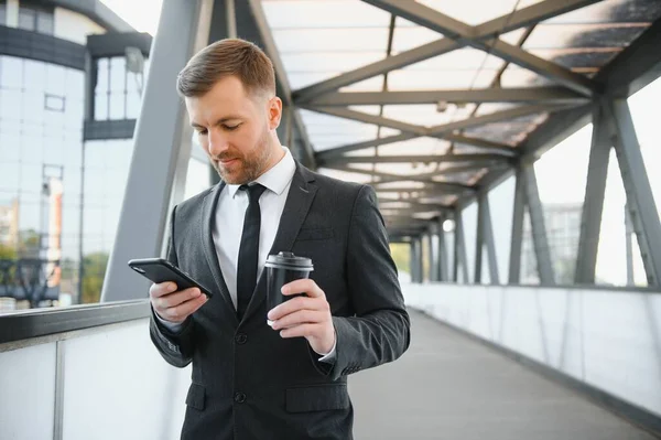 Bearded businessman in formal suit on break using mobile phone use smartphone. business man standing outside on modern urban city street background with coffee cup in downtown outdoors. copy space.