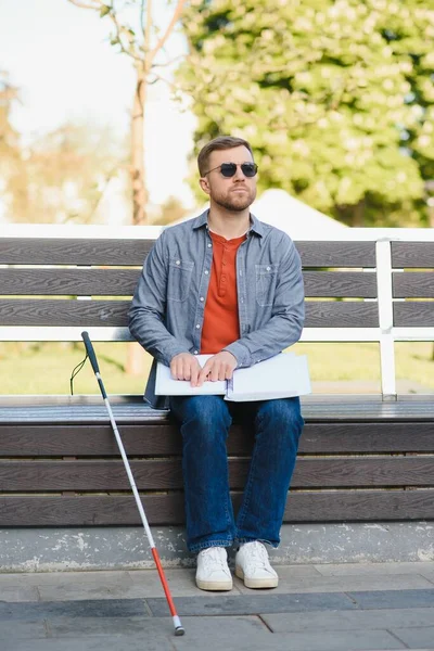 Blind man reading braille book, sitting on bench in summer park, resting.