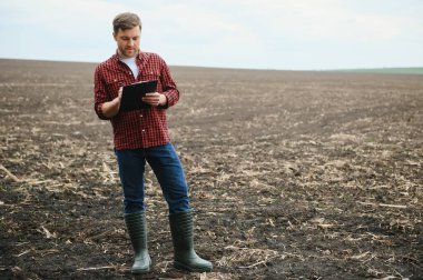 A farmer in boots works with his tablet in a field sown in spring. An agronomist walks the earth, assessing a plowed field in autumn. Agriculture. Smart farming technologies. clipart