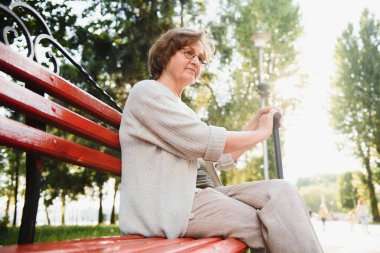 Tişörtlü zarif yaşlı kadın sıcak bir günde parkta bankta oturuyor..