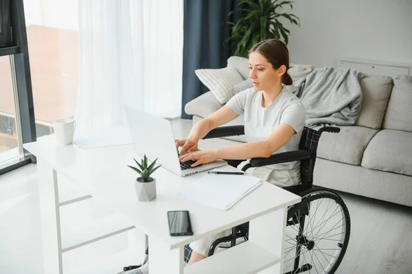 Freelancer in wheelchair using laptop near notebook and papers on table.