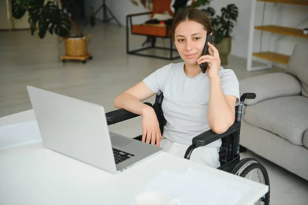 Freelancer in wheelchair using laptop near notebook and papers on table.