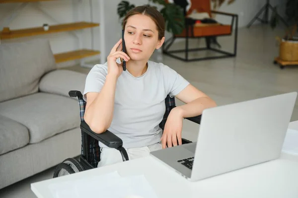 Freelancer in wheelchair using laptop near notebook and papers on table.