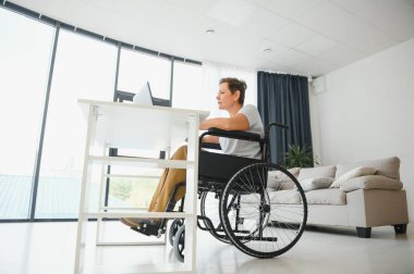 Tekerlekli sandalyedeki serbest yazar dizüstü bilgisayarını ve masadaki kağıtları kullanıyor..