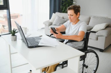 Orta yaşlı, gri saçlı, engelli bir kadın tekerlekli sandalyede otururken görüntülü konuşma yapıyor.