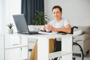Evde tekerlekli sandalyede oturan orta yaşlı kadın dizüstü bilgisayar kullanıyor..