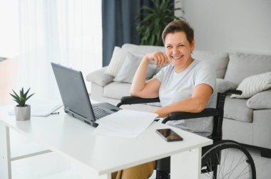 Tekerlekli sandalyedeki serbest yazar dizüstü bilgisayarını ve masadaki kağıtları kullanıyor..