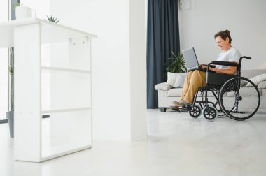 Evde tekerlekli sandalyede oturan orta yaşlı kadın dizüstü bilgisayar kullanıyor..