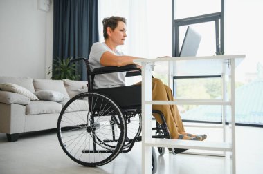 Evde tekerlekli sandalyede oturan orta yaşlı kadın dizüstü bilgisayar kullanıyor..