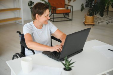 Tekerlekli sandalyedeki serbest yazar dizüstü bilgisayarını ve masadaki kağıtları kullanıyor..