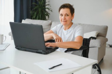 Evde tekerlekli sandalyede oturan orta yaşlı kadın dizüstü bilgisayar kullanıyor..
