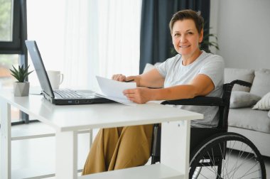 Tekerlekli sandalyedeki serbest yazar dizüstü bilgisayarını ve masadaki kağıtları kullanıyor..