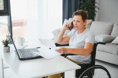 Evde tekerlekli sandalyede oturan orta yaşlı kadın dizüstü bilgisayar kullanıyor..