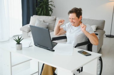 Tekerlekli sandalyedeki serbest yazar dizüstü bilgisayarını ve masadaki kağıtları kullanıyor..