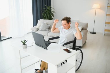 Tekerlekli sandalyedeki serbest yazar dizüstü bilgisayarını ve masadaki kağıtları kullanıyor..