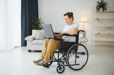 Evde tekerlekli sandalyede oturan orta yaşlı kadın dizüstü bilgisayar kullanıyor..