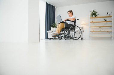 Evde tekerlekli sandalyede oturan orta yaşlı kadın dizüstü bilgisayar kullanıyor..