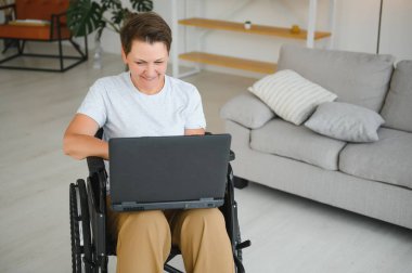 Evde tekerlekli sandalyede oturan orta yaşlı kadın dizüstü bilgisayar kullanıyor..