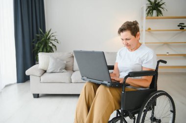 Evde tekerlekli sandalyede oturan orta yaşlı kadın dizüstü bilgisayar kullanıyor..