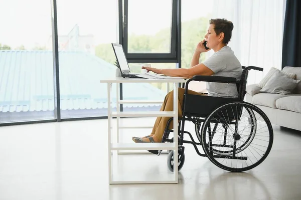 Freelancer in wheelchair using laptop near notebook and papers on table.